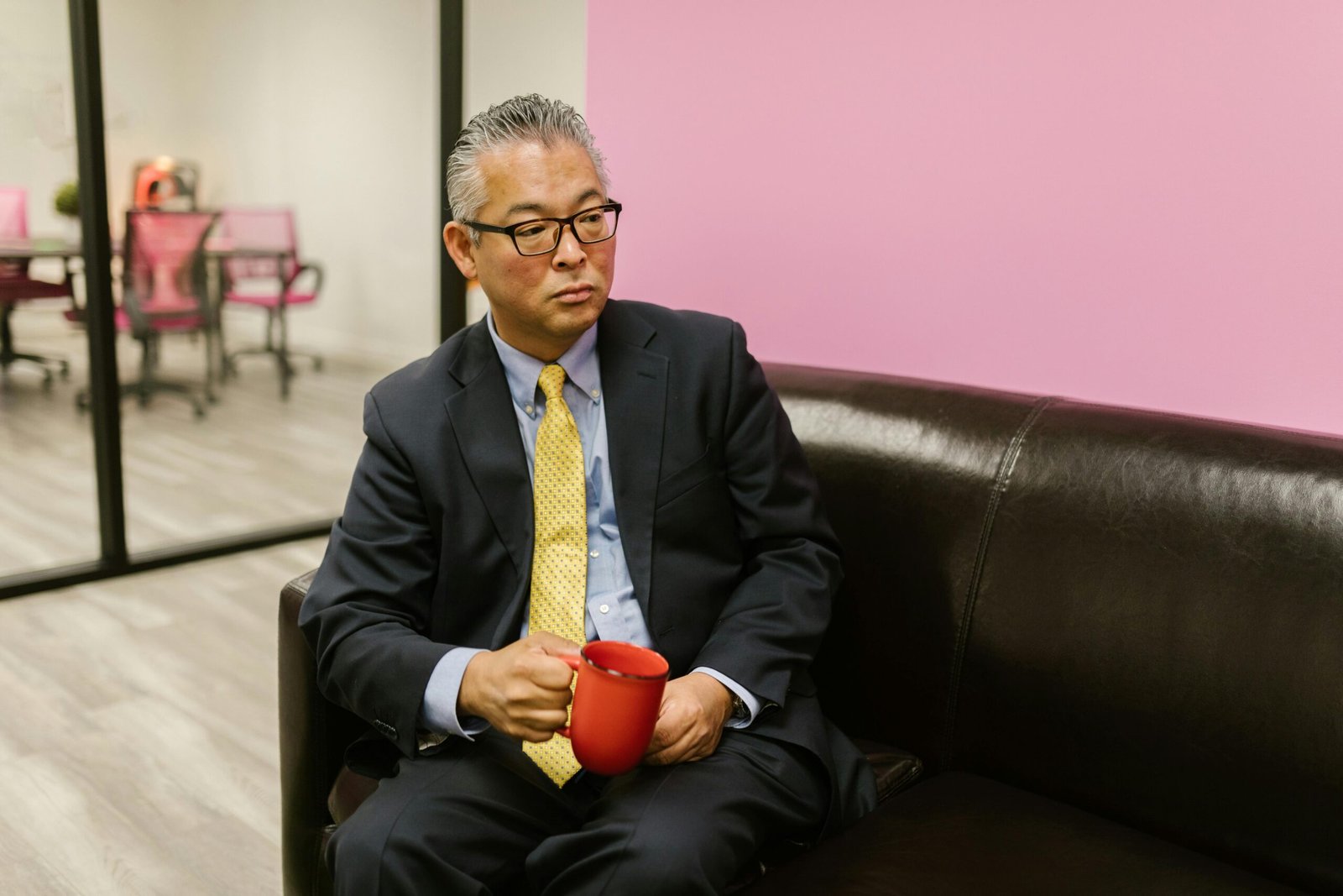 An Asian businessman in a suit, sitting on a sofa with a red mug in a modern office setting.